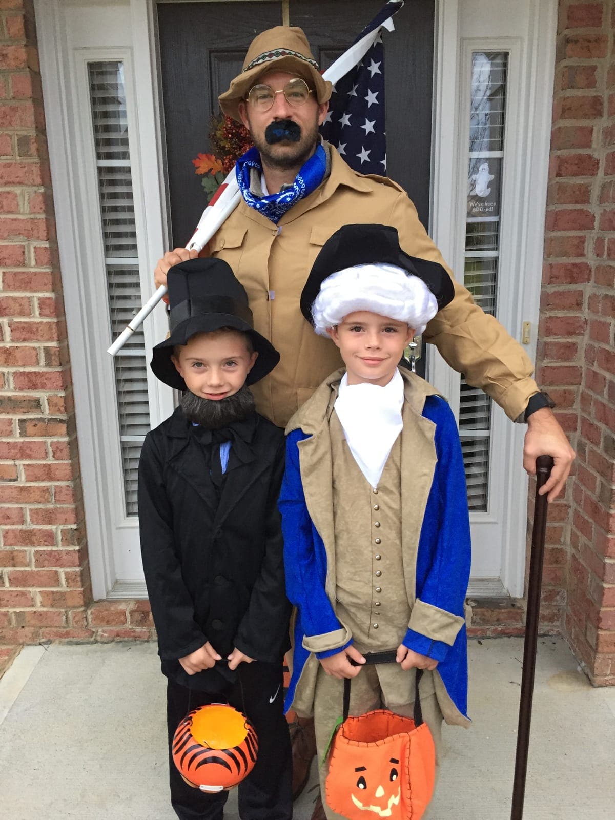 Mr. Joe dressed as Teddy Roosevelt with two boys dressed as Abraham Lincoln and George Washington on a front porch, holding trick-or-treat buckets.